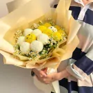 Bouquet of yellow and white ping pong chrysanthemums with smaller white flowers, wrapped in pale yellow paper.