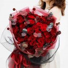 A bouquet of vibrant red roses with eucalyptus leaves, wrapped in pink and black paper.