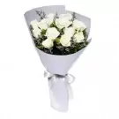A bouquet of white roses with delicate greenery, wrapped in light purple paper and tied with a white ribbon.