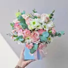 A hand holds a light blue box filled with pink and white roses, chrysanthemums, and eucalyptus leaves.