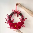 Red spring wreath with roses and berries, held by hands against a white wall.