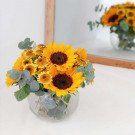 Glass bowl of sunflowers and greenery with reflection in mirror.