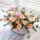 White basket with pink and orange roses, eucalyptus, and small blue flowers.