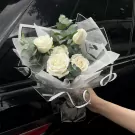 Bouquet of white roses with eucalyptus, wrapped in sheer white paper and black ribbon, held by a hand.