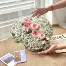 Pink roses and baby's breath arranged in a round bouquet on a burlap cloth, next to photos and a newspaper.