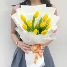 Woman holding a bouquet of 10 yellow tulips wrapped in white paper and tied with a peach ribbon.