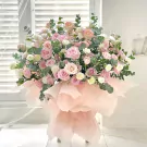 A large bouquet of pink roses, white carnations, and eucalyptus in a pink paper wrap.