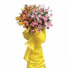 A large, bright floral arrangement featuring yellow orchids, pink roses, and greenery, adorned with yellow fabric.
