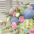 A bouquet of pink roses, blue chrysanthemums, and hydrangeas in a box with newspaper clippings.