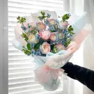 Rainbow roses and blue accent flowers in a bouquet, wrapped in pastel paper and tied with a pink ribbon.