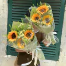 Two bouquets of sunflowers with smaller white flowers, wrapped in clear plastic and green paper, tied with white ribbon.
