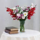 A bouquet of red orchids, purple mums, and pink lisianthus in a glass vase on a table with books.