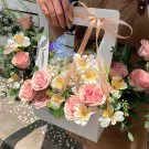 Pink roses, white and yellow flowers in a white basket with a blue butterfly and gold ribbon.