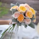 A bouquet of yellow, pink, and orange roses with dusty grey leaves and white ribbon.