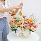 Hand holding lanterns over a flower basket with orange and pink roses, carnations, and daisies.