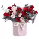 A pink hatbox filled with red and pink roses, greenery, and red berries, tied with a white ribbon.