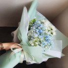 A bouquet of blue hydrangeas and white daisies wrapped in light green and white paper.