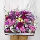 A wooden box arrangement of purple roses, carnations, orchids, and filler flowers, with gold-wrapped candies.
