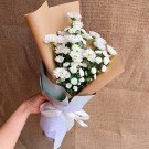 A hand holds a bouquet of white chrysanthemums and calimero flowers wrapped in brown paper and a blue accent with a white ribbon.