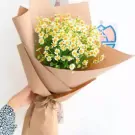 Hand holding a bouquet of small white daisies with yellow centers, wrapped in brown paper.