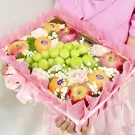 Fruit basket with green grapes, oranges, apples, and pink roses, wrapped in pink mesh.