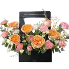 A floral arrangement in a black basket with peach roses, pink carnations, and daisies.