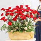 A large floral arrangement of red roses in a yellow basket, adorned with small white flowers and decorative rattan balls.