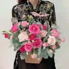 A basket of pink and red roses with greenery, held by a person in a floral top.