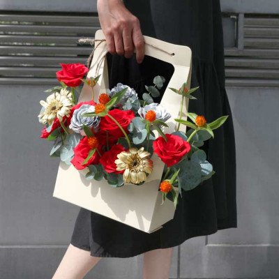 A woman holds a cream-colored flower basket filled with red roses, white gerbera daisies, and eucalyptus.