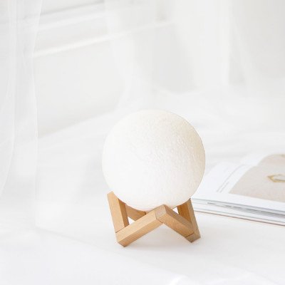 A white moon-shaped lamp with a textured surface sits on a wooden stand beside an open book.