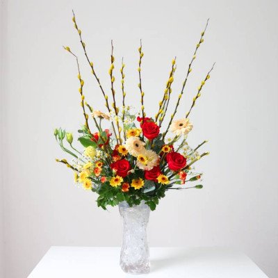 A tall floral arrangement with red roses, yellow and cream daisies, and yellow pussy willow branches in a clear vase.