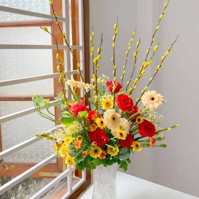 A vase of red roses, yellow sunflowers, gerbera daisies, and pussy willow branches.