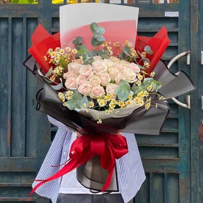 Bouquet of light pink roses, chamomile, and eucalyptus wrapped in black and red paper, tied with a red bow.