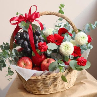 Fruit and flower basket with red gerbera daisies, white chrysanthemums, red carnations, red apples, and dark grapes.