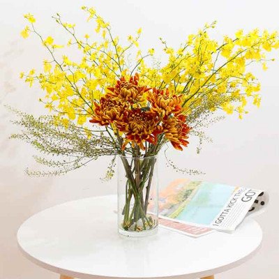 Yellow orchids and orange chrysanthemums in a glass vase on a white table.