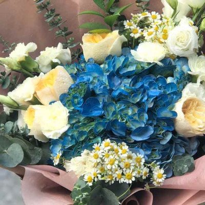 A bouquet of blue hydrangeas, white and yellow roses, and small white daisies.