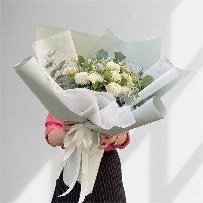 A person holding a pale green bouquet of white chrysanthemums and carnations, tied with a ribbon.