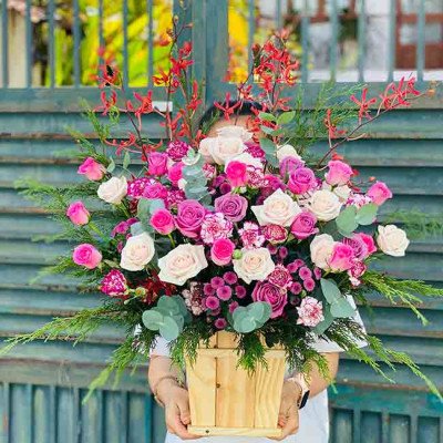 Wooden box overflowing with pink and white roses, carnations, and greenery, held by hands.
