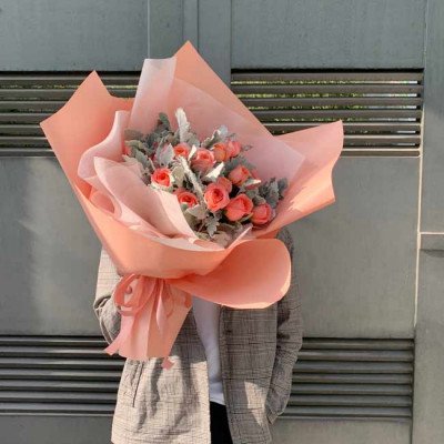 A bouquet of peach roses and grey leaves wrapped in peach and white paper.