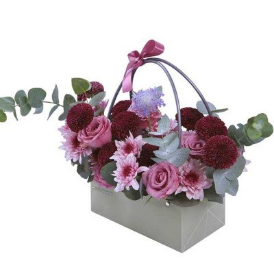 A bouquet of pink roses, purple flowers, and green foliage in a grey gift box with a pink ribbon.