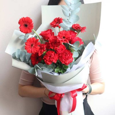 Red roses, gerberas, and carnations in a bouquet with eucalyptus leaves, wrapped in paper and tied with a red bow.