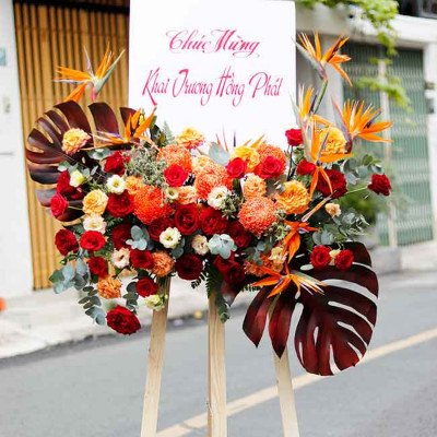 Flower stand with red and orange roses, Birds of Paradise, and large tropical leaves, bearing Vietnamese congratulatory text.