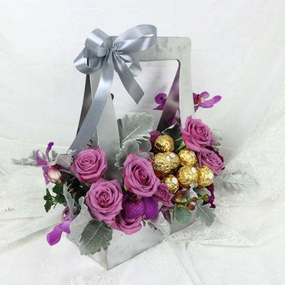 A gift box with purple roses, Ferrero chocolates, and orchids, tied with a silver ribbon.