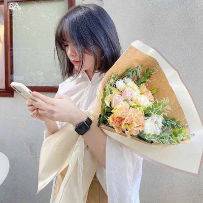 A woman in a white shirt holds a pastel flower bouquet, looking at her phone.