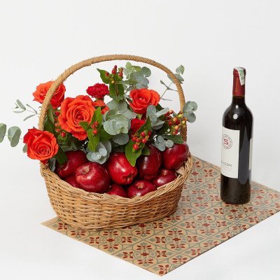Wicker basket overflowing with red apples, orange roses, and greenery, next to a bottle of red wine.