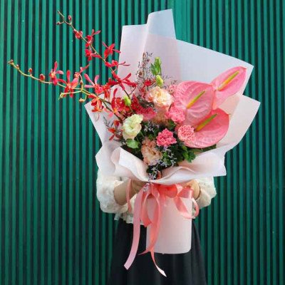 A bouquet of pink anthuriums, red orchids, and carnations wrapped in pink paper.