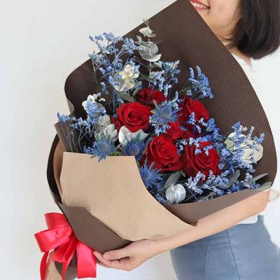 Bouquet of red Ecuador roses with blue statice and thistle, wrapped in brown and tan paper, tied with a red bow.