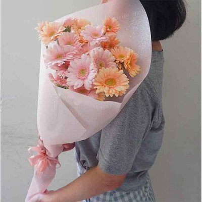 A bouquet of pink and orange gerbera daisies wrapped in light pink paper, held by a person in a grey shirt.