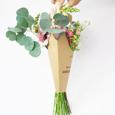 A hand holds a bouquet of spring flowers in a brown paper cone holder.