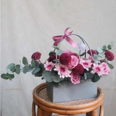 Pink and purple floral arrangement in a grey box with a pink ribbon on a wicker stool.